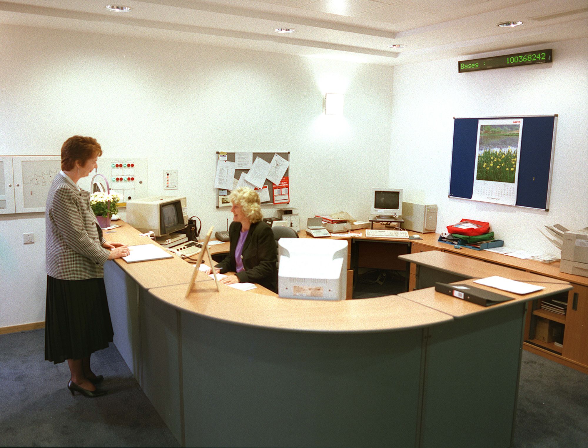 Two women are chatting on either side of a reception desk, one standing and one sitting. Behind them, a digital counter with the word Bases displays the number: 100368242