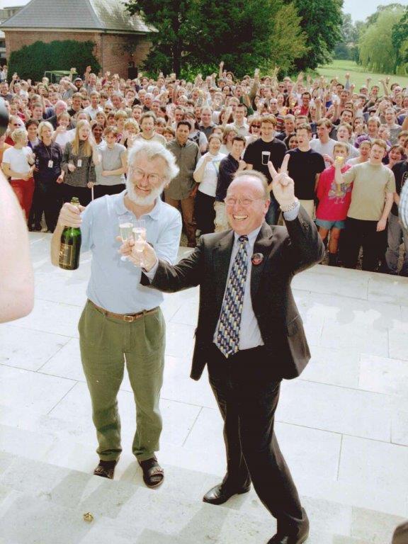 At the front of the image, two men hold up glasses of champagne and grin at a photographer whose arm can be seen. They are standing in front of a cheering crowd, some of whom are also holding up drinks.