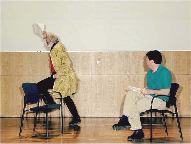 On the left of the image, a man wearing a long golden coat leaps onto a chair holding a sheet of paper aloft in his right hand. Next to him, a man is sitting looking at him and smiling, legs crossed and also holding a sheet of paper.