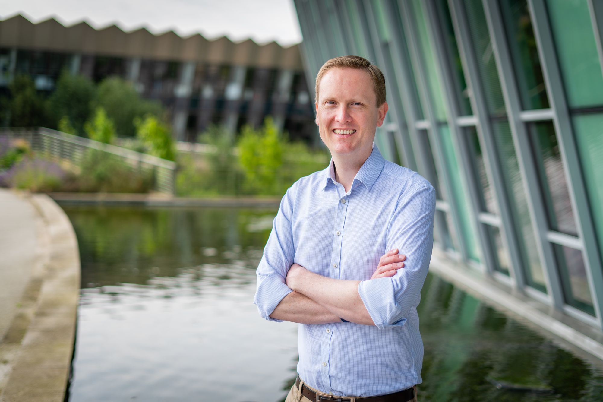 A white man with short hair smiles at the camera. Facing away from the viewer, he stands with his arms crossed in front of a shallow pool at the entrance to a glass-fronted building. Green plants and another building can be seen in the background.