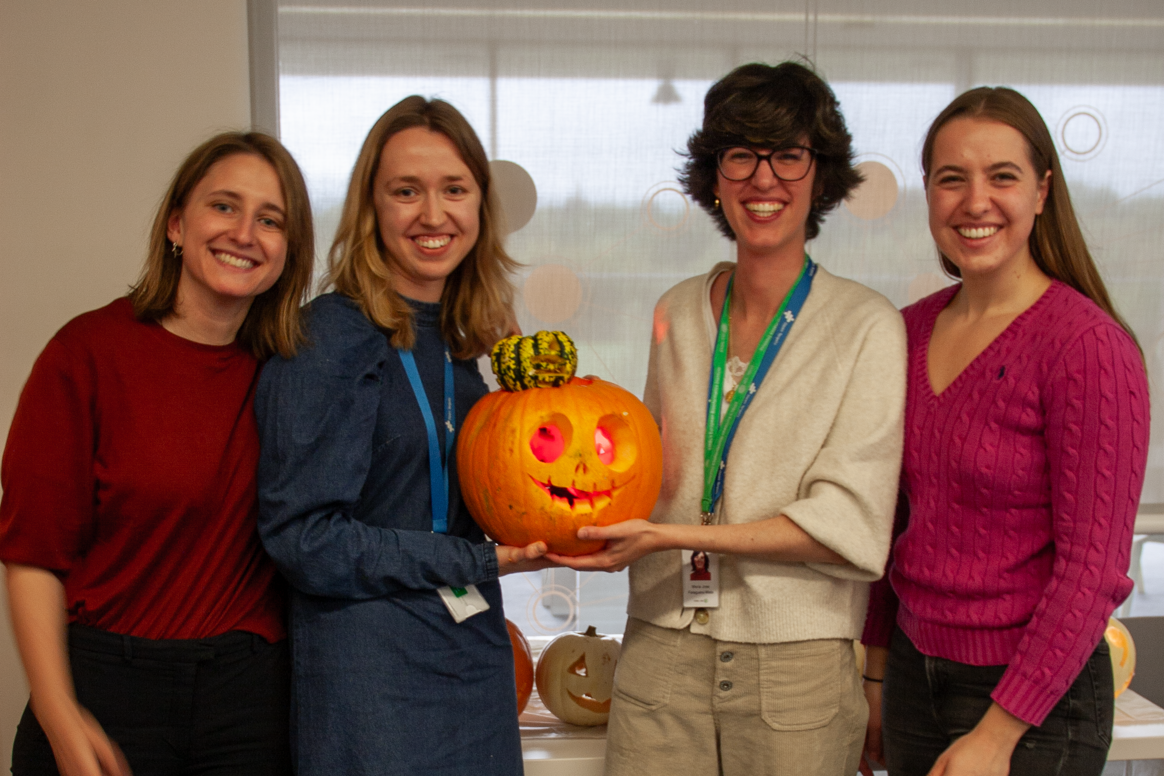 Four women stand in a line, smiling at the camera. They are holding up a carved orange pumpkin with a crooked smile, on top of which sits a small green pumpkin carved with the Open Targets logo. More pumpkins can be seen on the table behind them.