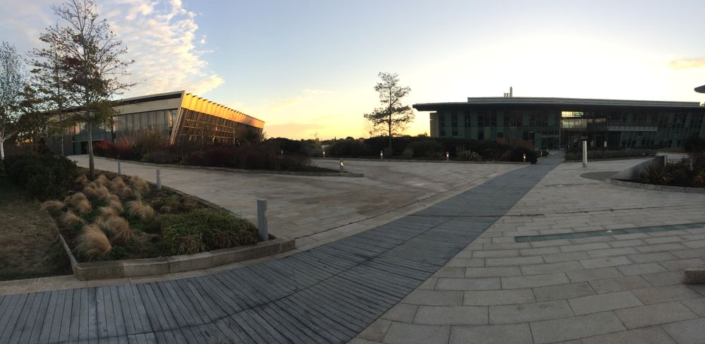 Panoramic view of the entrance to the Campus: a walkway leads up to two buildings, one of which reflects the setting sun.